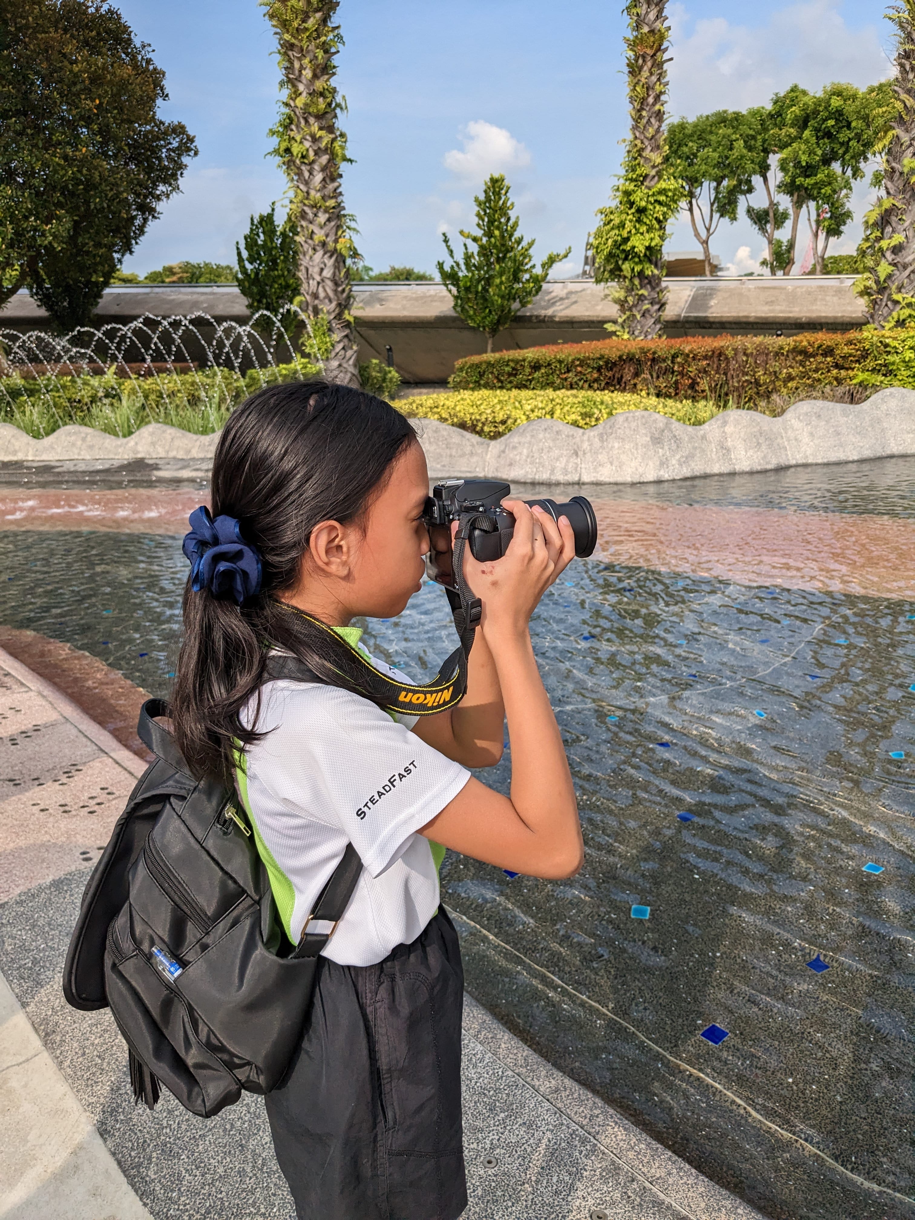 Girl with ponytail and backpack taking a photo by a shallow fountain pool, palms and hedges behind.